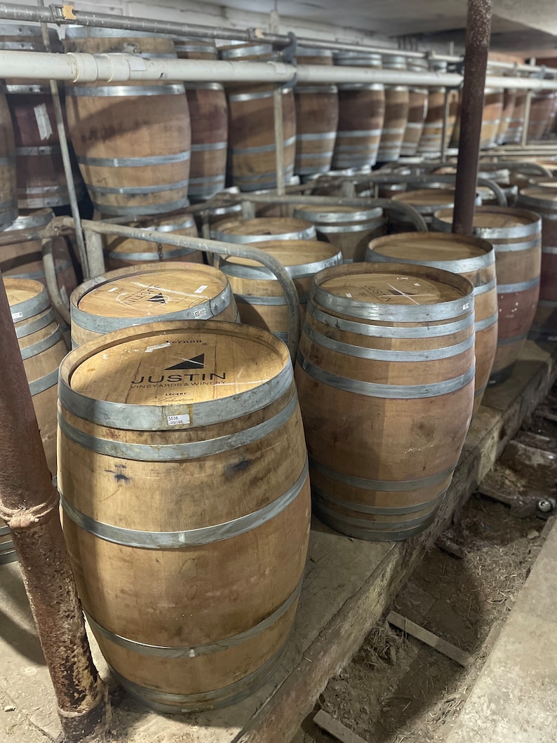 May include: A row of wooden barrels stacked in a winery. The barrels are made of oak and have metal bands around them. The barrels are labelled with the name "Justin Vineyards & Winery".