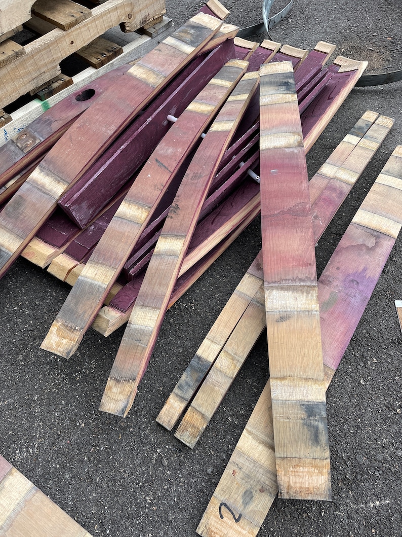 May include: A pile of reclaimed wine barrel staves, some are stained a deep purple, others are natural wood colour. The staves are arranged in a random pattern, some are curved, others are straight. The staves are all about 2.5 cm wide and 90 cm long.