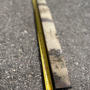 May include: A weathered, rectangular piece of wood, approximately 36 inches long, lies next to a yellow measuring tape. The tape shows inch markings. The wood has dark, aged markings and a rough texture, suggesting it's reclaimed or repurposed.