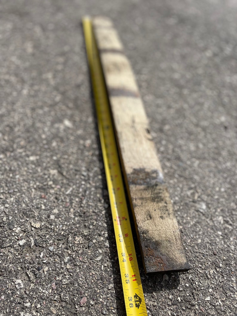 May include: A weathered, rectangular piece of wood, approximately 36 inches long, lies next to a yellow measuring tape. The tape shows inch markings. The wood has dark, aged markings and a rough texture, suggesting it's reclaimed or repurposed.