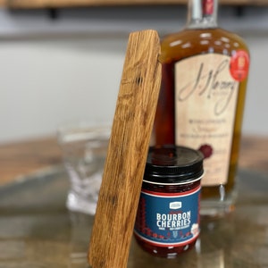 May include: A wooden bottle stopper with a natural wood grain finish. The stopper is shaped like a rectangle with rounded edges. It is sitting on a wooden surface next to a bottle of bourbon and a jar of bourbon cherries.