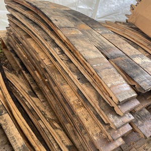 May include: A stack of reclaimed, weathered wooden barrel staves. The staves are curved, showing the original shape of the barrel, and display a mix of light and dark brown tones with areas of black. The wood has a rustic, aged appearance.