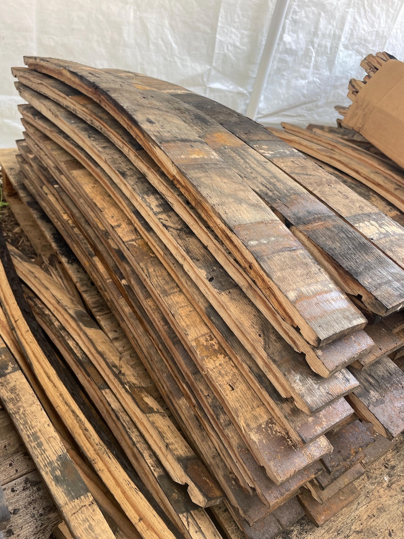 May include: A stack of reclaimed, weathered wooden barrel staves. The staves are curved, showing the original shape of the barrel, and display a mix of light and dark brown tones with areas of black. The wood has a rustic, aged appearance.
