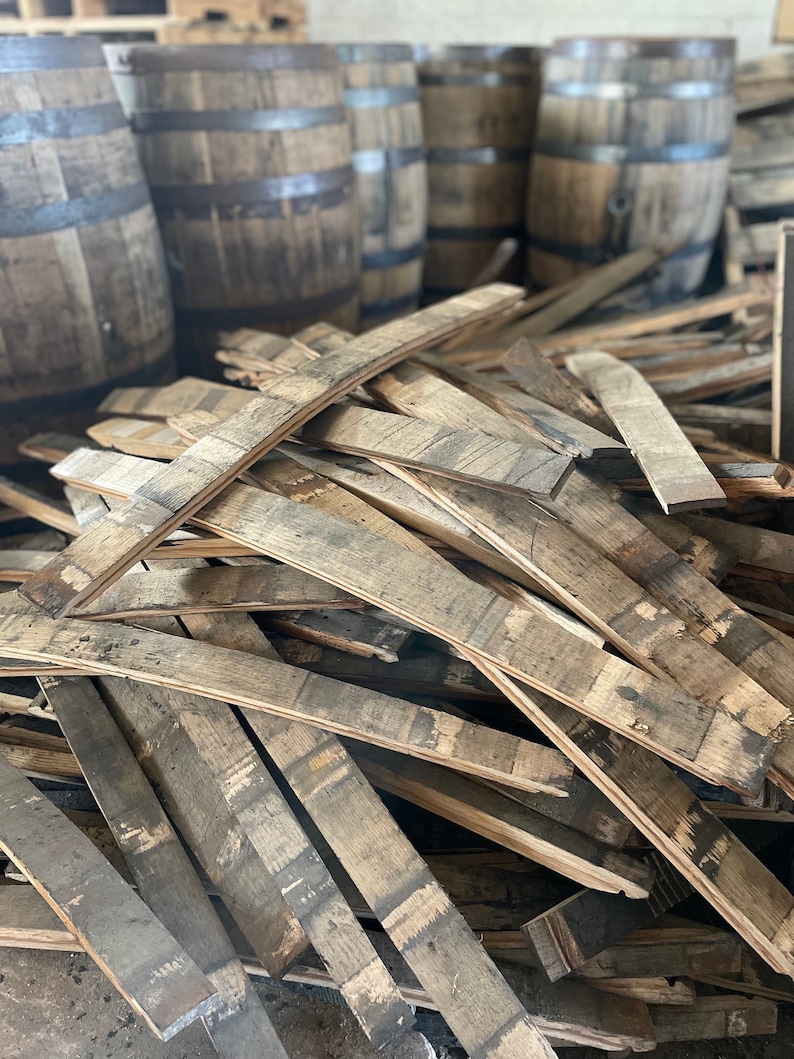 May include: A pile of reclaimed wooden barrel staves, showing the natural grain and weathered appearance. Several wooden barrels with metal bands are in the background, suggesting a distillery or aging process. The wood has a mix of light and dark brown tones.