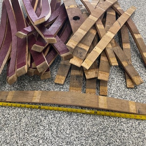 May include: A pile of wooden barrel staves, some are stained a deep burgundy red and others are a natural wood colour. A yellow measuring tape is laid out on the floor next to the staves.