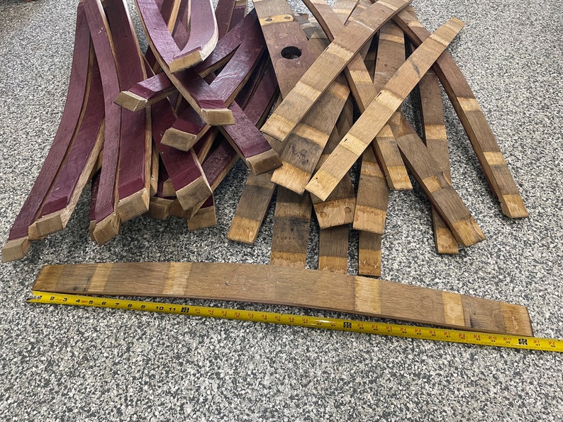 May include: A pile of wooden barrel staves, some are stained a deep burgundy red and others are a natural wood colour. A yellow measuring tape is laid out on the floor next to the staves.