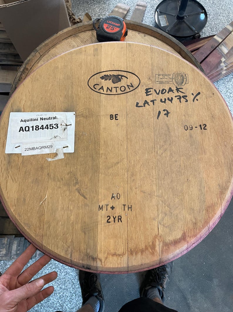 May include: A wooden barrel with the text "CANTON" and "EVOAK LAT 4475% 17" written on it. The barrel is labelled with the text "Aquilini Neutral AQ184453 22MBAQRM29" and "BE 40 MT TH 2YR".