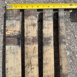May include: Four wooden planks, each approximately 28 cm long, with a dark brown stain and a light brown wood grain. The planks are arranged side by side on a grey asphalt surface.