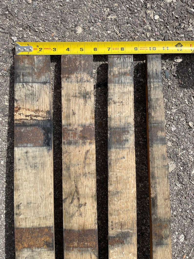 May include: Four wooden planks, each approximately 28 cm long, with a dark brown stain and a light brown wood grain. The planks are arranged side by side on a grey asphalt surface.