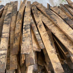 May include: A pile of reclaimed, weathered wooden barrel staves. The planks are various lengths and widths, showing dark charring and staining. The wood has a rustic, aged appearance, with visible grain and texture.