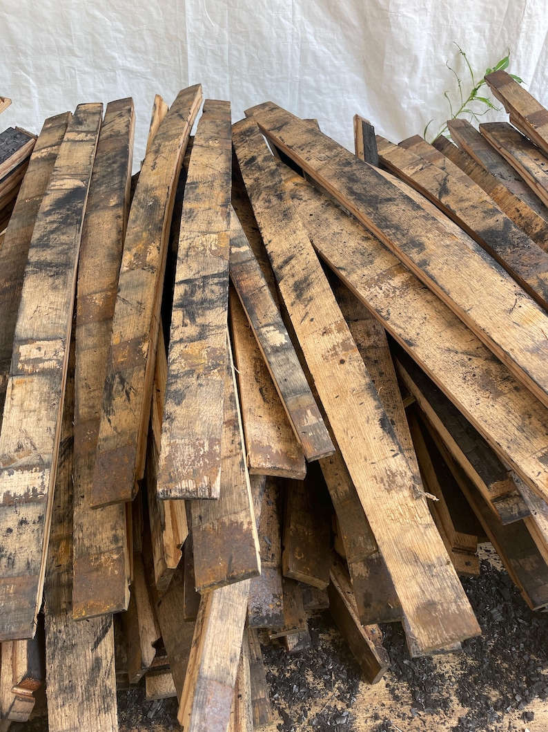 May include: A pile of reclaimed, weathered wooden barrel staves. The planks are various lengths and widths, showing dark charring and staining. The wood has a rustic, aged appearance, with visible grain and texture.