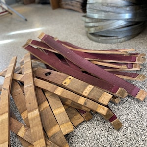 May include: A pile of wooden barrel staves, some are natural wood colour and some are stained a deep burgundy red. The staves are stacked in a random pattern and are ready to be used for making barrels.