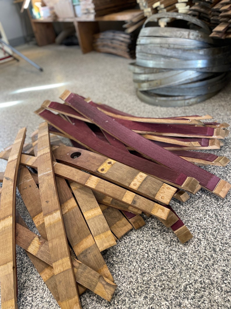 May include: A pile of wooden barrel staves, some are natural wood colour and some are stained a deep burgundy red. The staves are stacked in a random pattern and are ready to be used for making barrels.