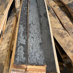 May include: Close-up of charred oak barrel staves. The interior of the staves is deeply blackened and textured, while the exterior shows a lighter, weathered wood grain. The staves are arranged in a pile, showcasing the raw, rustic appearance.