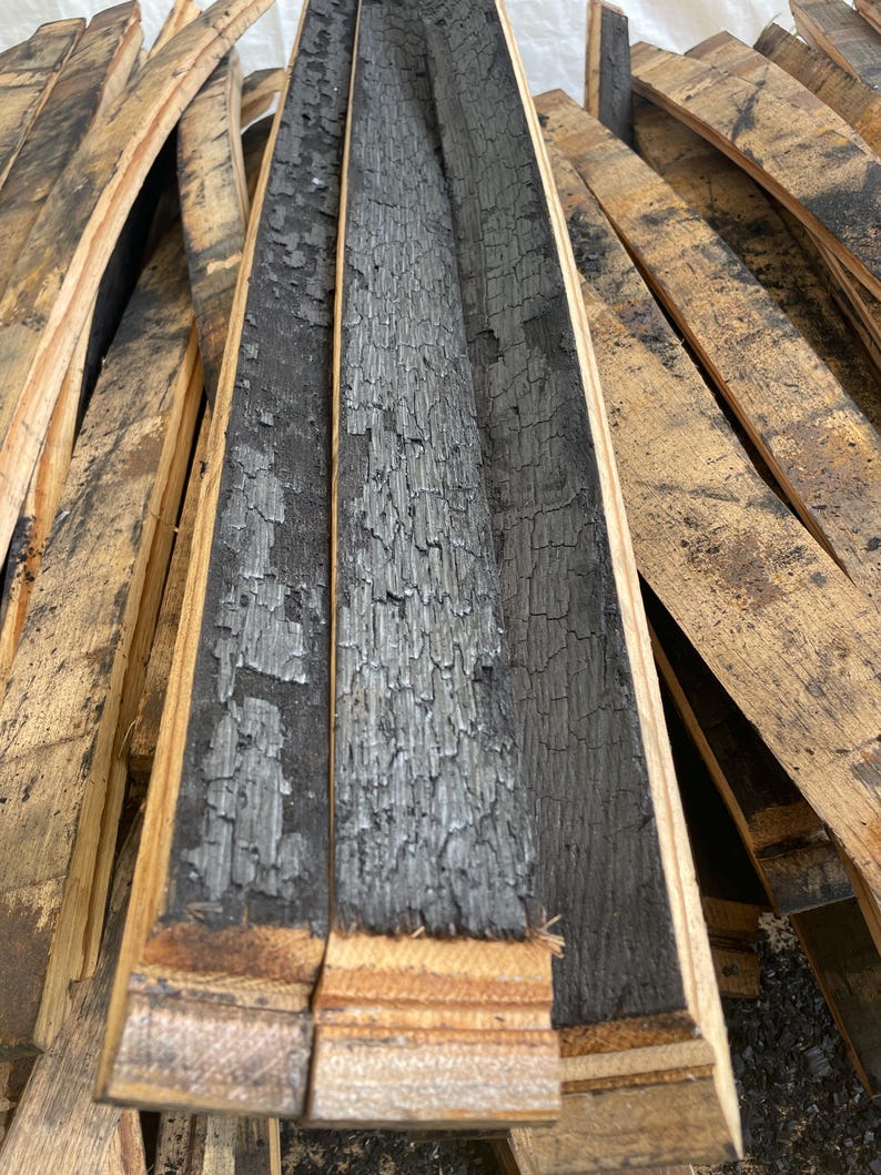 May include: Close-up of charred oak barrel staves. The interior of the staves is deeply blackened and textured, while the exterior shows a lighter, weathered wood grain. The staves are arranged in a pile, showcasing the raw, rustic appearance.