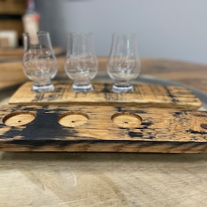 Może przedstawiać: Dwa drewniane tace do degustacji whisky z trzema uchwytami na kieliszki każda. Tace wykonane są z odzyskanych beczek po bourbonie i mają ciemnobrązowe wykończenie.
