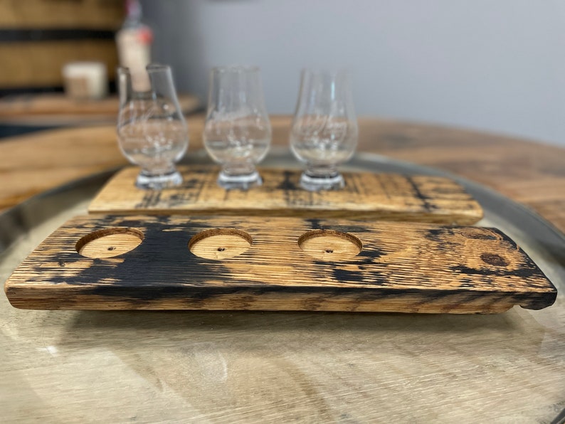 Może przedstawiać: Dwa drewniane tace do degustacji whisky z trzema uchwytami na kieliszki każda. Tace wykonane są z odzyskanych beczek po bourbonie i mają ciemnobrązowe wykończenie.