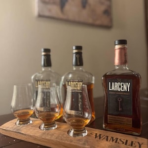 Może przedstawiać: Trzy butelki Larceny Straight Bourbon Whiskey, dwie ze szkła przezroczystego i jedna z miedzianą butelką, na drewnianej tacy z trzema szklankami wypełnionymi bursztynowym płynem. Taca jest oznaczona etykietą "Wamsley".