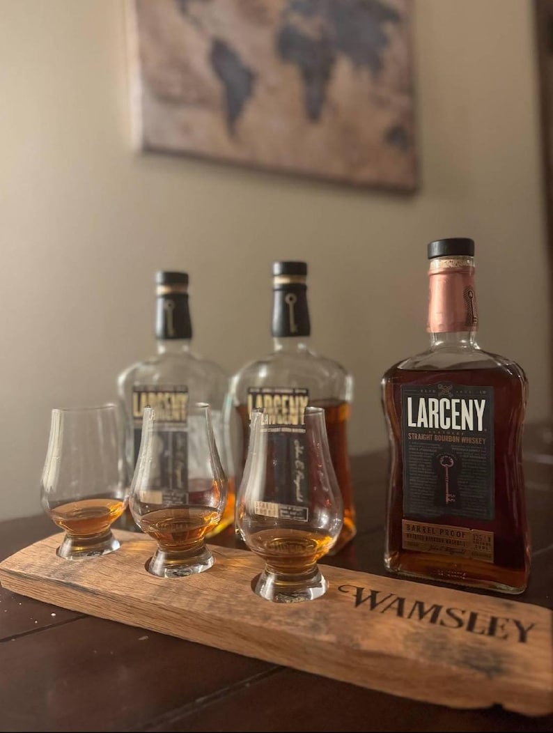 Może przedstawiać: Trzy butelki Larceny Straight Bourbon Whiskey, dwie ze szkła przezroczystego i jedna z miedzianą butelką, na drewnianej tacy z trzema szklankami wypełnionymi bursztynowym płynem. Taca jest oznaczona etykietą "Wamsley".