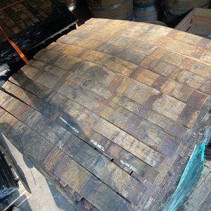 May include: A stack of reclaimed wooden barrel staves, showing the aged, weathered appearance of the wood. The staves are held together with metal bands, and are ready for reuse in woodworking projects. The wood has a mix of brown and gray tones.