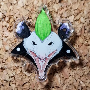May include: A clear acrylic pin with a cartoon image of a possum with a green mohawk, black and white fur, and a toothy grin.