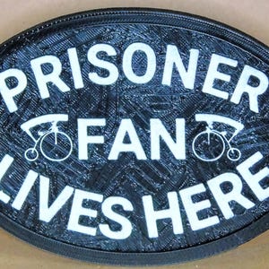 Könnte beinhalten: Ein schwarzes ovales Schild mit weißem Text, der "Prisoner of Fan Lives Here" lautet. Das Schild hat zwei kleine Fahrradbilder auf beiden Seiten des Wortes "Fan".