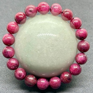 Op de afbeelding: Een cirkelvormige armband bestaande uit gepolijste, bolvormige kralen in een diepe magenta-roze tint. Elke kraal vertoont natuurlijke variaties in kleurintensiteit en textuur, wat zorgt voor een consistente en levendige uitstraling.