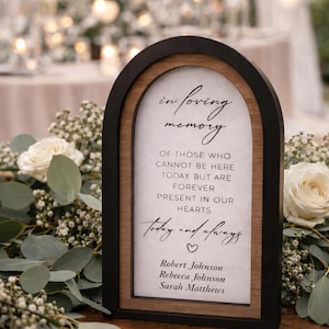 May include: A wooden memorial sign with a black frame and a light wood interior. The sign reads "In loving memory" and includes names. The sign is surrounded by white flowers and greenery, suitable for a wedding or memorial.