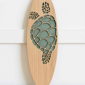 May include: Wooden surfboard-shaped wall art featuring a laser-cut sea turtle design. The turtle's shell is filled with a teal-colored background. The surfboard is made of light-colored wood and is mounted on a white wall.