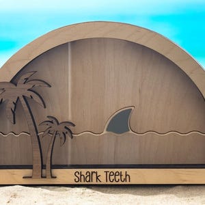 May include: Wooden beach-themed display with a shark fin, palm trees, and the text "Shark Teeth." The design is cut out of light-colored wood, with a dark border. The background is a blurred beach scene.