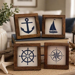 Könnte beinhalten: Vier quadratische Holzrahmen mit dunkelbraunem Finish, die jeweils einen weißen Einsatz mit einem dunkelblauen nautischen Motiv enthalten: ein Anker, ein Segelboot, ein Steuerrad und eine Windrose. Die Rahmen sind in einem Zweierraster angeordnet.