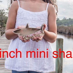 A person holds a small wooden shark-shaped shadow box with a white outline and the words "Shark Teeth" engraved on the base, standing on a wooden dock.