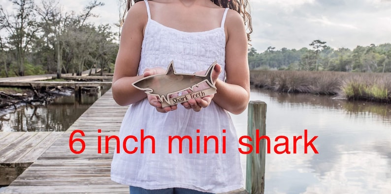 A person holds a small wooden shark-shaped shadow box with a white outline and the words "Shark Teeth" engraved on the base, standing on a wooden dock.