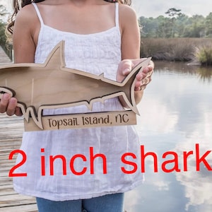 A person holds a larger wooden shark-shaped shadow box with "Topsail Island, NC" engraved on its side, standing on a wooden dock.