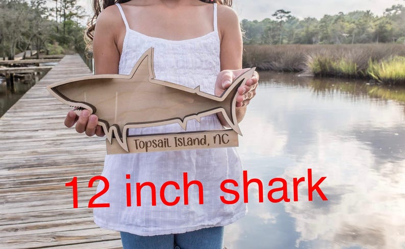 A person holds a larger wooden shark-shaped shadow box with "Topsail Island, NC" engraved on its side, standing on a wooden dock.