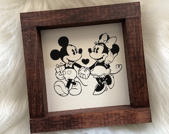 May include: A framed sign featuring a black and white illustration of Mickey and Minnie Mouse holding hands, with a heart between them. Mickey Mouse is holding a gift. The frame is made of dark brown wood.
