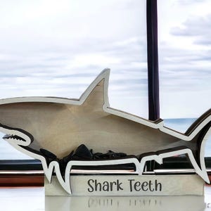 A wooden shark-shaped shadow box with a white outline and the words "Shark Teeth" engraved on the base, containing small dark objects.