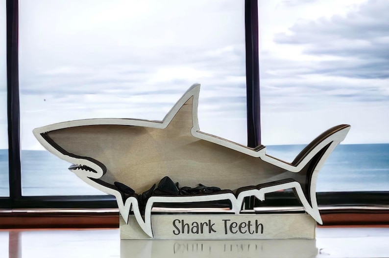 A wooden shark-shaped shadow box with a white outline and the words "Shark Teeth" engraved on the base, containing small dark objects.