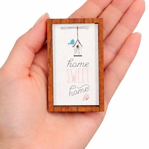 以下が含まれることがあります： 茶色のフレームが付いた、小さな長方形の木製サイン。サインには、鳥小屋と青い鳥のイラスト、そして手書き風の「home SWEET home」という言葉が描かれています。ピンク色の蝶も描かれています。