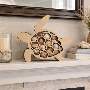 Peut inclure: Étagère en bois en forme de tortue remplie de coquillages. L'étagère est marron clair et présente un motif en nid d'abeille. Une bougie dans un bocal en verre, un corail décoratif et des livres sont également sur l'étagère.