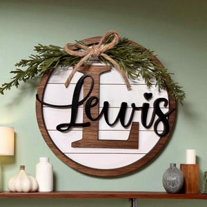 Puede incluir: Un letrero redondo de madera con el nombre "Lewis" en escritura negra. El letrero tiene un fondo blanco con listones de madera horizontales, un marco de madera marrón y está decorado con vegetación y un lazo de cuerda. Un corazón está encima de la "i".