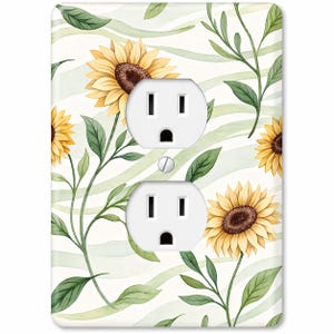 Puede incluir: Cubierta de enchufe eléctrica blanca con un diseño de acuarela de girasoles y hojas verdes sobre un fondo a rayas verdes claras y blancas. La cubierta tiene dos enchufes eléctricos estándar.