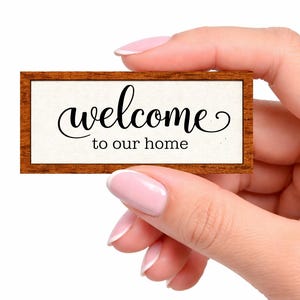 May include: A rustic wooden sign with the text "Welcome to our home" in white lettering. The sign features a distressed finish and a simple, inviting design, perfect for a front porch or entryway.
