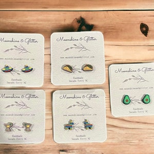 May include: Six pairs of handmade earrings on white cards with the text "Moonshine & Glitter" and a website address. The earrings feature various designs, including a sombrero, a taco, an avocado, a margarita glass, and a pinata.