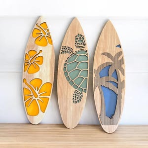 May include: Three wooden surfboards with decorative cutouts. One features yellow hibiscus flowers, another a teal turtle, and the last a palm tree with a blue background. The surfboards are displayed against a white wall.