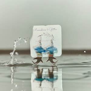 May include: A pair of handmade resin earrings featuring a blue and yellow ocean wave design with silver hooks. The earrings are displayed on a white card with the text "Moonshine & Glitter" and "www.moonshineandglitter.com".