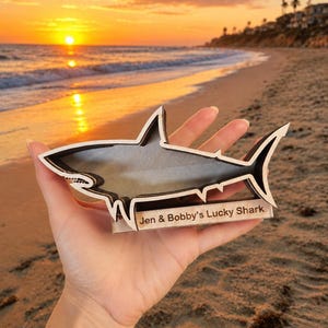 A hand holds a wooden shark-shaped shadow box with "Jen & Bobby's Lucky Shark" engraved on it, silhouetted against a sunset beach scene.