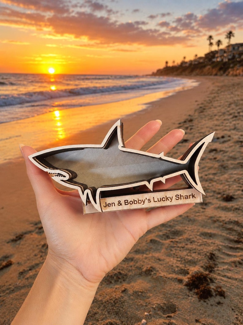 A hand holds a wooden shark-shaped shadow box with "Jen & Bobby's Lucky Shark" engraved on it, silhouetted against a sunset beach scene.