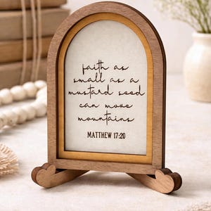 May include: Wooden arched picture frame with the quote "faith as small as a mustard seed can move mountains" from Matthew 17:20. The frame is light brown with a cream-colored background and a decorative base.