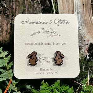 May include: A pair of wooden bunny stud earrings with white accents. The earrings are displayed on a white card with the text "Moonshine & Glitter" and "www.moonshineandglitter.com". The card also includes the text "Handmade" and "Sneads Ferry NC".